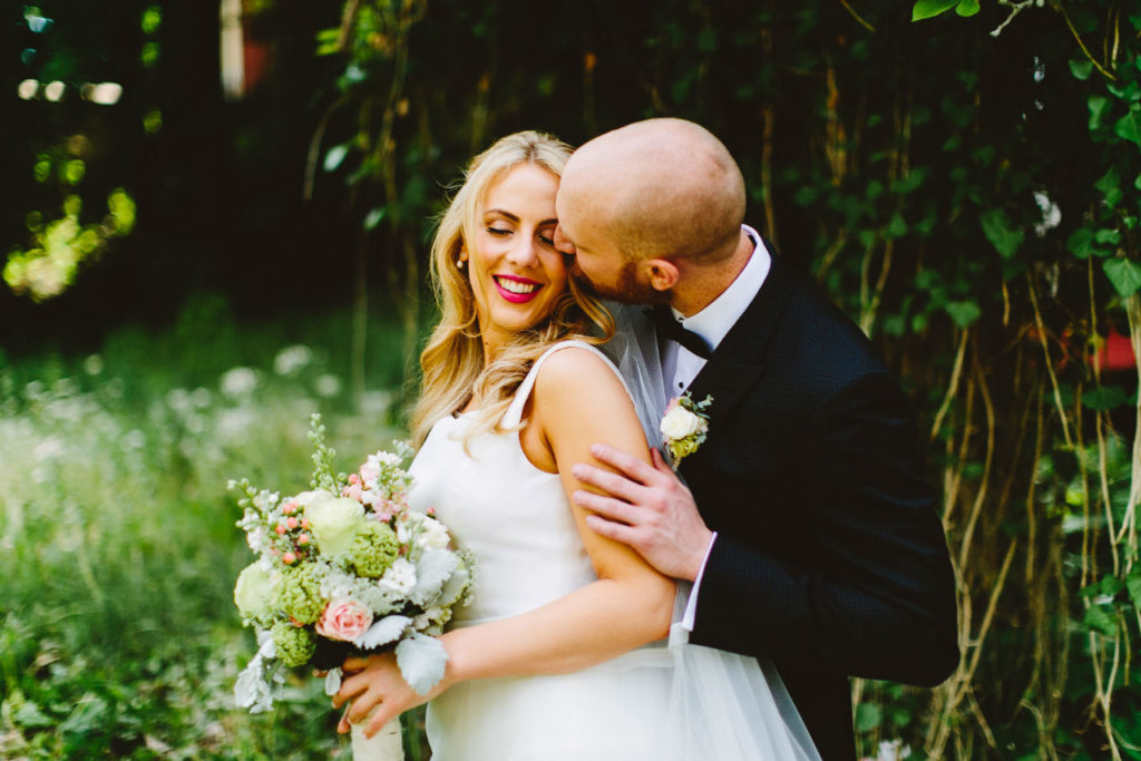 Groom kissing smiling bride holding bouquet outdoors